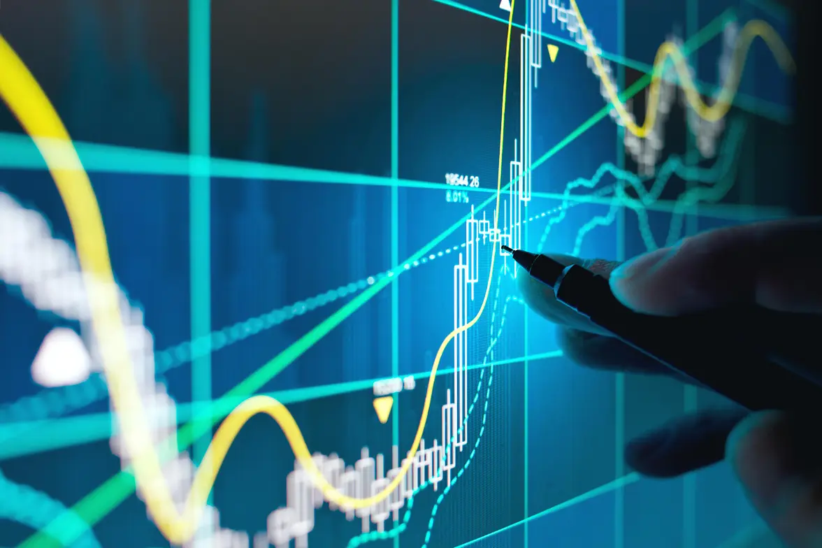 A hand holding a pen pointing at a screen displaying financial graphs and percentages.