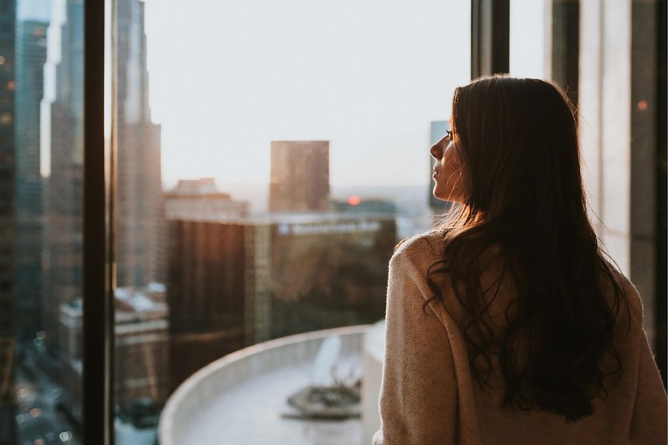 Woman gazing out a high-rise window at a cityscape bathed in the warm glow of sunrise, contemplating the urban scene.