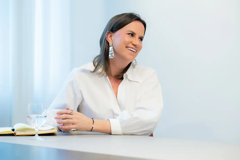 A young, dark-haired woman in a white blouse smiles openly at another person who is apparently sitting at a table with her.